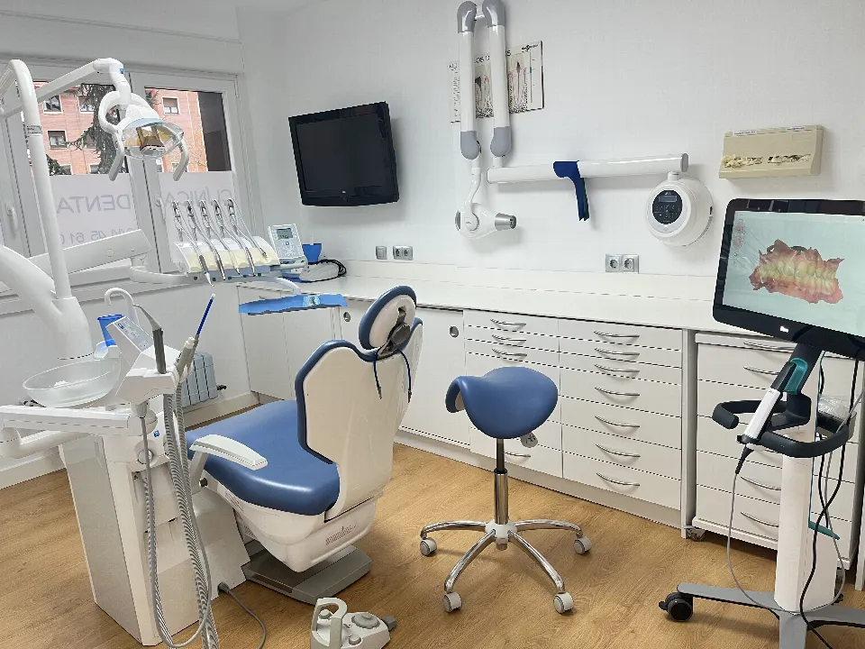 Un gabinete, con el sillón azul, el taburete, el aparato de rayos en la pared y el escáner intraoral con su pantalla a la derecha
